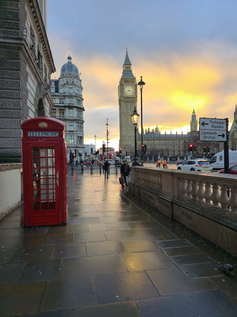 A street scene in London during sunset shows a classic red telephone booth positioned on the paved sidewalk close to a historic building with ornate architecture. In the background, the iconic Big Ben clock tower and the Palace of Westminster are visible under a partly cloudy sky with warm hues of orange and yellow. Street lamps illuminate the area, and a few pedestrians are walking along the pavement, some carrying bags. On the right side, a stone railing separates the sidewalk from the road where cars are passing by. A directional street sign points towards Trafalgar Square and Lambeth Bridge. The scene suggests an urban environment, typical of central London, with nearby traffic and foot traffic indicating active movement related to house removals or relocation services. Lambeth Man and Van may be involved in transportation and packing tasks within this context of moving logistics.