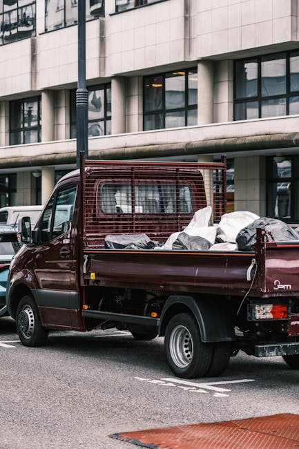 A dark red flatbed truck parked on a city street during daylight hours, loaded with various household items prepared for removal. The truck's cargo area contains multiple large, wrapped furniture pieces covered with protective blankets and secured with straps, along with several cardboard boxes and plastic bags filled with household belongings. The vehicle is positioned close to a modern office building with large windows and a concrete exterior, indicating an urban environment suitable for home relocation services. The truck is fitted with a metal mesh side panel to contain the load, and a towing hitch is visible at the rear. Nearby, there's a white van and other vehicles, with a lamppost and pavement in view, supporting the scene of ongoing furniture transport and packing during the moving process, consistent with professional removals services like those offered by Lambeth Man and Van.