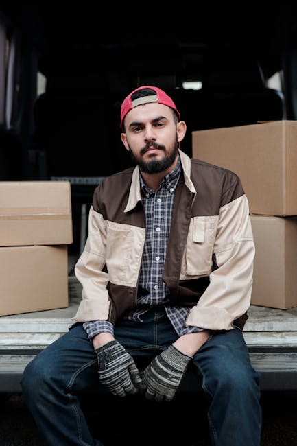 A young man with a beard, wearing a pink baseball cap backwards, a checkered shirt, and a beige and brown jacket, is sitting on the edge of a van's loading area with a neutral expression. Surrounding him are several closed cardboard boxes, some piled on the vehicle's floor, indicating a home relocation or furniture transport process. The interior of the van is dark, with the back doors open, revealing a black headrest of a seat behind him. The scene suggests a professional removals service, such as Lambeth Man and Van, preparing for a moving or packing and moving operation involving careful handling of household items, with the man possibly taking a break during the loading process.