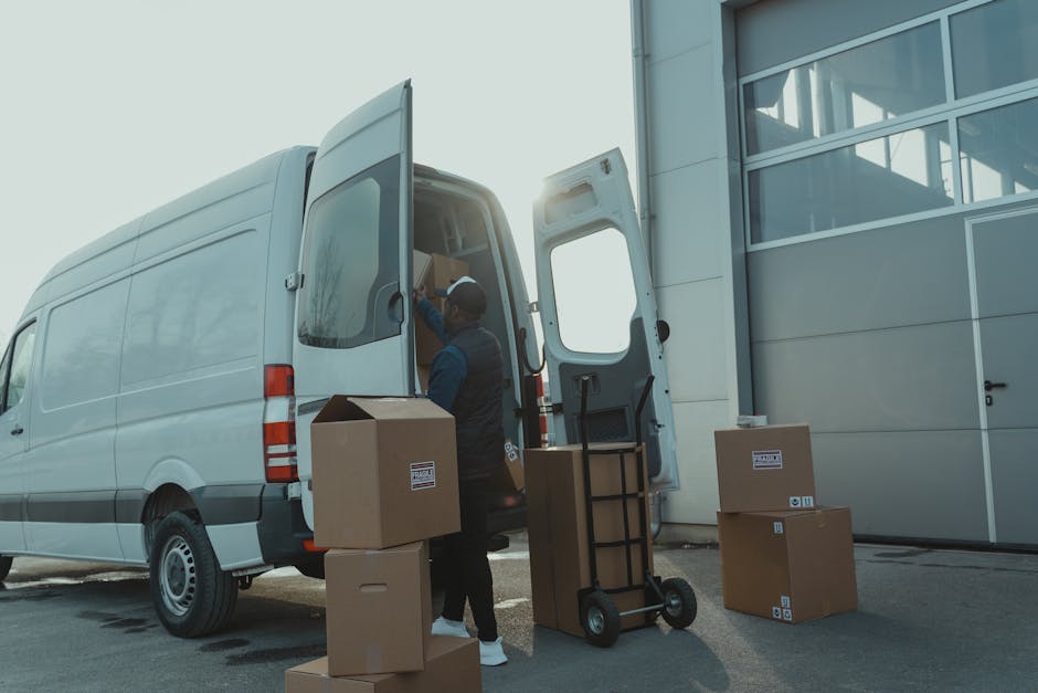 A professional mover from Lambeth Man and Van is loading a white delivery van parked outside a modern building with large glass windows on a clear day. The mover, wearing a dark jacket and cap, is seen reaching into the van’s interior, which contains packed cardboard boxes of various sizes, some sealed with packing tape, and wrapped furniture. Several additional cardboard boxes are placed on the ground nearby, awaiting transport. A black hand trolley with a metal frame and rubber wheels stands next to the van, ready to assist with furniture transport and packing and moving procedures. The loading process takes place on pavement adjacent to the building, with the van door wide open, illuminated by natural daylight, highlighting the logistics involved in house relocations and flat access solutions, as provided by Lambeth Man and Van's removals services.