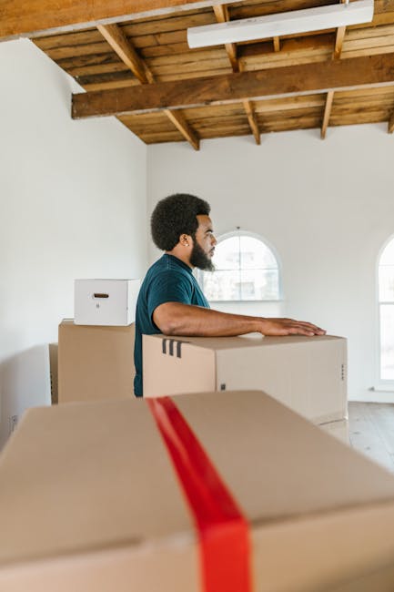 A young man with a beard, wearing a pink baseball cap backwards, a checkered shirt, and a beige and brown jacket, is sitting on the edge of a van's loading area with a neutral expression. Surrounding him are several closed cardboard boxes, some piled on the vehicle's floor, indicating a home relocation or furniture transport process. The interior of the van is dark, with the back doors open, revealing a black headrest of a seat behind him. The scene suggests a professional removals service, such as Lambeth Man and Van, preparing for a moving or packing and moving operation involving careful handling of household items, with the man possibly taking a break during the loading process.