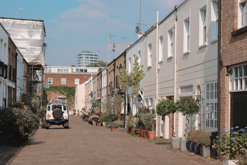 A narrow street in Clapham Common lined with terraced houses featuring white and brick facades, with front gardens decorated with potted plants and small trees. The street is paved with cobblestones and has a white van parked on the left side, likely involved in home relocation or furniture transport. The van is positioned near the curb, with rear doors potentially open for loading or unloading furniture and packed boxes wrapped in plastic or cardboard. Nearby, a dolly or trolley may be used for moving large items, and blankets or straps could be visible, indicating careful protection of belongings during the loading process. Residential buildings have multiple windows, some with barred designs, and the scene is illuminated by natural daylight, reflecting the typical logistics involved in house removals through narrow streets and steps in Lambeth. Lambeth Man and Van's professional services are implied in the context of the moving process depicted.