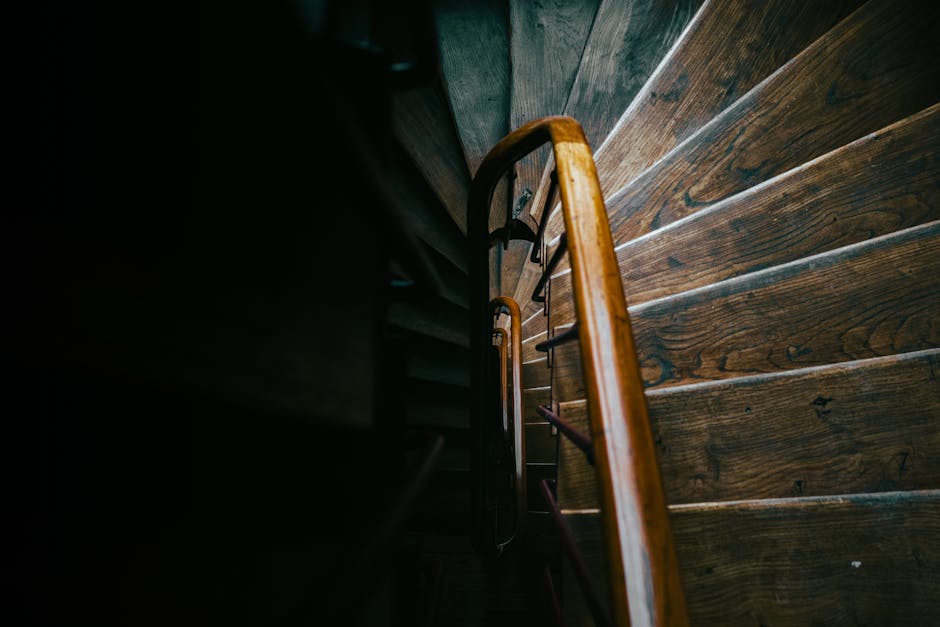 Top-down view of a narrow wooden staircase inside a property during a house move, with a polished wooden handrail and dark metal balustrades visible. The staircase is illuminated by natural light, hig