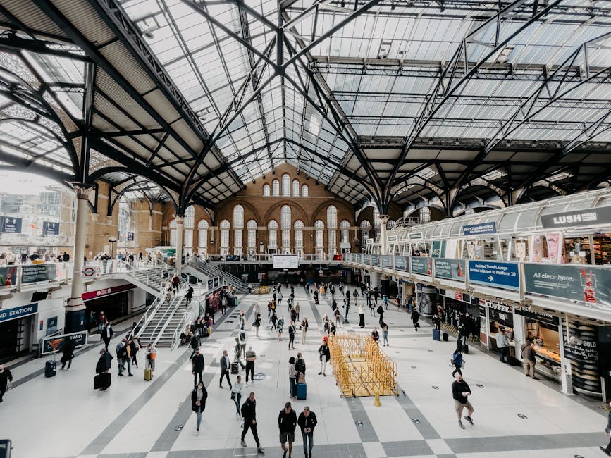 Interior view of a large train station with a high, arched glass and metal ceiling supported by black metal framework. The spacious concourse is filled with numerous travellers, some walking with lugg