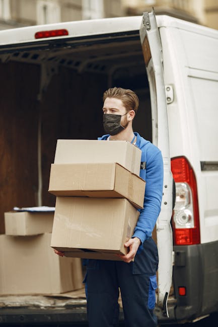 A man wearing a black face mask and dressed in a blue hoodie and dark trousers is standing outside the open rear doors of a white moving van. He is holding three cardboard boxes stacked together, with the top box being larger and the bottom a smaller one. Inside the van, additional cardboard boxes and packing materials such as paper or bubble wrap are visible, some placed on the floor and others stacked against the interior walls. The scene takes place on a street with a building facade seen in the background. The lighting suggests daytime, and the overall setting reflects a home relocation or furniture transport process, with the individual in the act of loading or unloading items as part of a move coordinated by Lambeth Man and Van, a professional removals service. This image illustrates the packing and loading stage of house removals, emphasizing careful handling of packed boxes and organized vehicle loading for efficient house moving operations.
