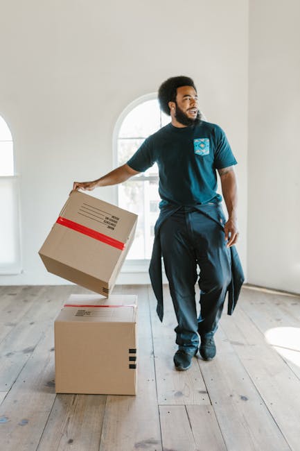 A professional mover from Lambeth Man and Van is inside an empty, well-lit room with white walls and light wooden flooring, during a house removal process. The mover, a man with a dark complexion and curly black hair, is wearing a navy blue t-shirt with a small logo on the chest and dark trousers, with a jacket tied around his waist. He is holding a cardboard box wrapped with red packing tape in his right hand, preparing to place it on a stack of two other cardboard boxes, one on the floor and one being lowered from above. The boxes are standard moving cartons, some with printed labels, and are positioned near an arched window that allows natural light to illuminate the space. Behind him, the room appears empty, indicating a packing or loading stage of a home relocation. The environment suggests careful handling of packing materials, likely in preparation for transport or furniture transport as part of a moving logistics operation executed by Lambeth Man and Van, a professional removals company. The scene emphasizes tasks associated with packing and loading during a same-day, last-minute move, with focus on the careful handling of boxes in a residential setting.