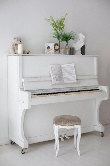 A white upright piano with an open sheet of music resting on its music stand, situated indoors against a light gray wall. The piano is on small castor wheels, facilitating easy movement, and accompanied by a matching small, upholstered stool with a fabric seat. On top of the piano, there are decorative items including a small teddy bear, a candle, and framed photographs, alongside a clear glass vase containing green foliage. To the right, there is a white bust sculpture and a dark container, all positioned on top of the piano, which appears to be in a room prepared for a house relocation or packing and moving process. The overall setting is well-lit, emphasizing the clean, minimalistic aesthetic suitable for professional removals involving furniture transport and home relocation services, consistent with the offerings of Lambeth Man and Van.