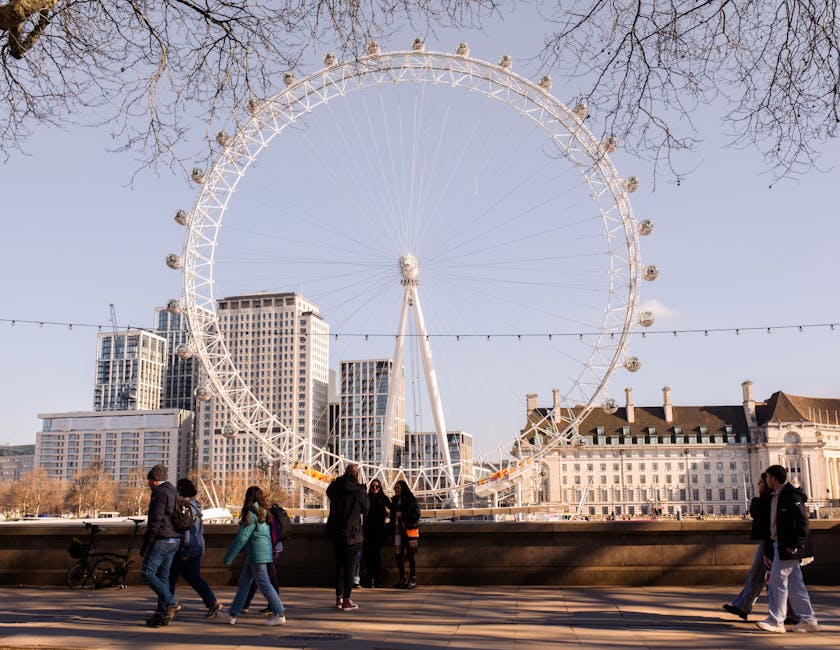 A large, white observation wheel, known as the London Eye, dominates the skyline of London on a clear day with a blue sky. Several leafless trees frame the top of the image, indicating it may be autumn or winter. In the foreground, a paved walkway along the River Thames features a group of pedestrians walking past, some of whom are engaged in conversation, while others are looking at the wheel. To the right, a few individuals are walking alone or in pairs, dressed in casual clothing, suitable for cool weather. Along the riverbank, a low stone wall separates the walkway from the water, with some bicycles parked nearby. The scene captures the vibrant urban environment typical of South Bank, with modern high-rise buildings in the background. This image is relevant for house removals and moving services related to city relocations, transport, and community logistics, showcasing the busy, outdoor setting where moving activities might take place, with Lambeth Man and Van often providing associated services.