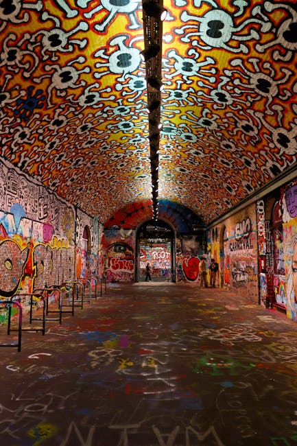 A long, arched underground passageway with a curved ceiling and walls covered in vibrant graffiti art, including various tags, symbols, and colorful illustrations. The ceiling features a pattern of white skulls with red outlines against a multicolored background, and a black pipe runs along the center of the ceiling, with a suspended chain or cable attached to it. The floor is also decorated with graffiti, paint splatters, and markings. On the left side, a row of metal benches is visible, while on the right side, a few individuals are seen walking or standing near the graffiti-covered walls. Natural light filters through the open exit at the far end of the tunnel, providing some illumination. The scene suggests an urban environment that may be part of a larger project involving interior decoration, street art, or possibly a designated area for artistic expression, relevant for house removals or furniture transport involved in relocation services, as managed by Lambeth Man and Van.