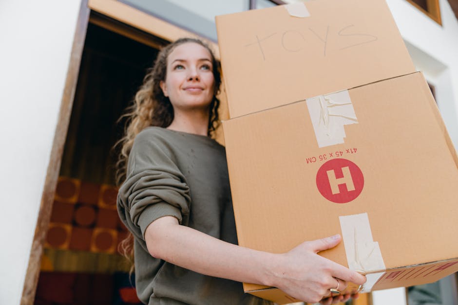 A young woman with curly hair and light skin is inside a home, holding two cardboard boxes stacked one on top of the other, preparing for a house move. The boxes are sealed with packing tape, with one labelled 'toys' handwritten on the top. She is wearing a casual, long-sleeved grey sweatshirt. The background shows an open doorway leading to another room with wooden flooring and stacked logs visible, indicating a inside residential environment. The lighting is natural and soft, highlighting her calm expression and the contents of the boxes. This scene exemplifies the packing process involved in home relocation, as part of furniture transport and moving logistics, supported by professional removal services like Lambeth Man and Van.