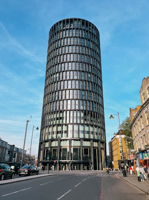 A tall, modern office building with a cylindrical shape and numerous vertical glass windows covering its facade, situated on a busy city street. The ground level features large glass entrance doors, and the surrounding area includes parked cars, pedestrians, and streetlights. In the foreground, a moving van part of Lambeth Man and Van's fleet is parked near the sidewalk, ready for a house relocation or furniture transport. Visible moving equipment such as trolleys, blankets, and boxes may be used to load household or office items onto the van as part of packing and moving services. This setting highlights the urban environment where professional removals are conducted, supporting efficient relocation logistics in Lambeth, possibly involving loading and transport processes for small business or residential moves.
