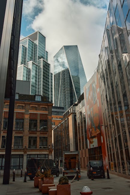 A daytime street scene showing part of a modern city centre with tall glass skyscrapers reflecting the sky and surrounding buildings. In the foreground, there are parked black vans and a small transport truck, some of which are used for home relocation or furniture transport, positioned near a commercial building with large windows and a patterned facade. Adjacent to this, older brick buildings with multiple stories are visible, creating a contrast between architectural styles. The pavement area features potted plants and some traffic cones, with a few bollards separating the loading zone from pedestrian pathways. The overall scene depicts an urban environment suitable for house removals, highlighting the typical loading and unloading area used during professional moving services such as those provided by Lambeth Man and Van for office and residential relocations in Lambeth.