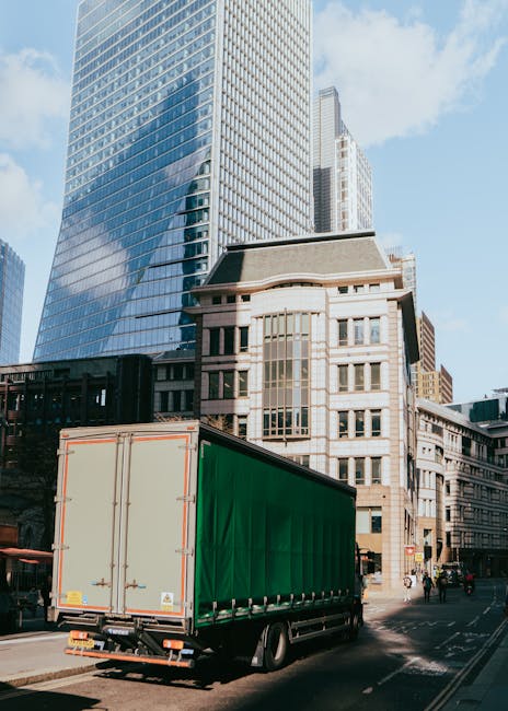 A tall, modern office building with a cylindrical shape and numerous vertical glass windows covering its facade, situated on a busy city street. The ground level features large glass entrance doors, and the surrounding area includes parked cars, pedestrians, and streetlights. In the foreground, a moving van part of Lambeth Man and Van's fleet is parked near the sidewalk, ready for a house relocation or furniture transport. Visible moving equipment such as trolleys, blankets, and boxes may be used to load household or office items onto the van as part of packing and moving services. This setting highlights the urban environment where professional removals are conducted, supporting efficient relocation logistics in Lambeth, possibly involving loading and transport processes for small business or residential moves.
